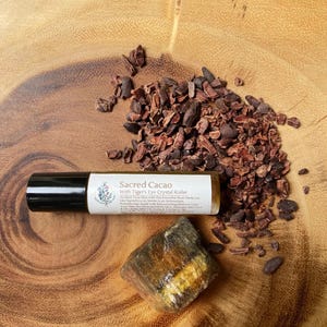 May include: A roll-on bottle labeled "Sacred Cacao With Tiger's Eye Crystal Roller" next to a piece of tiger's eye crystal and scattered cocoa nibs. The bottle has a black cap and a white label with text and a floral design. The items are on a wooden surface.