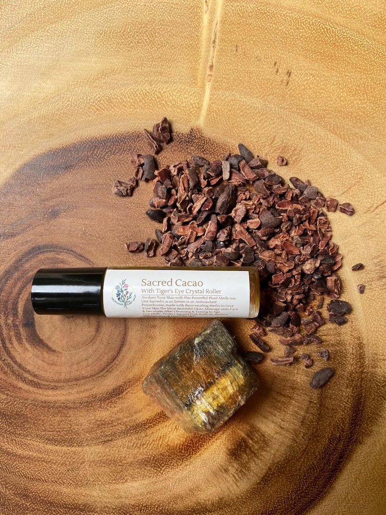 May include: A roll-on bottle labeled "Sacred Cacao With Tiger's Eye Crystal Roller" next to a piece of tiger's eye crystal and scattered cocoa nibs. The bottle has a black cap and a white label with text and a floral design. The items are on a wooden surface.