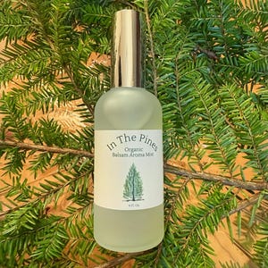 May include: A frosted glass bottle with a gold-colored cap, labeled "In The Pines Organic Balsam Aroma Mist" with a tree graphic. The bottle contains a light green liquid and is set against a backdrop of green pine branches. The bottle is 4 fl oz.