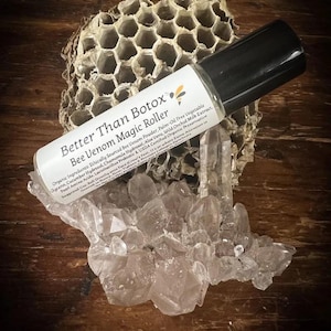 May include: A black-capped roller bottle labeled "Better Than Botox Bee Venom Magic Roller" rests on a cluster of clear crystals. The background includes a piece of honeycomb and a wooden surface. The label lists organic ingredients.