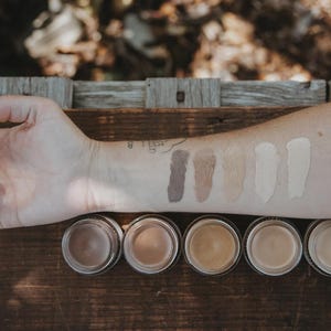 May include: An arm displaying foundation swatches and jars, showcasing a range of shades from dark brown to light beige. The swatches are applied to the skin, with matching jars below. The jars and arm are on a wooden surface.