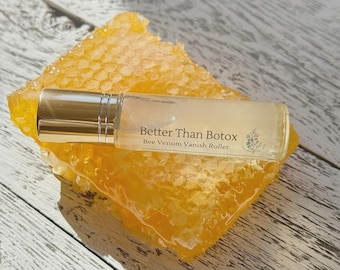 Könnte beinhalten: Eine transparente Glas-Rollerflasche mit goldenem Verschluss, beschriftet mit "Better Than Botox Bee Venom Vanish Roller", liegt auf einem Stück Honigwabe. Die Honigwabe ist leuchtend gelb und hat eine strukturierte Oberfläche. Der Hintergrund ist eine helle Holzoberfläche.