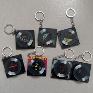 Peut inclure: Plusieurs porte-clés sur le thème des disques vinyles. Chaque porte-clés présente un design de disque miniature avec une pochette d'album, fixé à un support carré noir et un porte-clés argenté. Les designs varient.