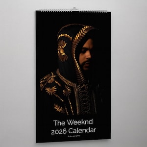 Pode incluir: Um calendário preto de 2026 com um retrato de The Weeknd. A imagem mostra The Weeknd usando uma roupa preta com capuz e bordados dourados. O calendário está pendurado em uma parede branca.