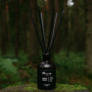 Op de afbeelding: Een zwarte glazen diffuserfles met zwarte stokjes, met het label "Cedar Wood". De fles staat op een met mos bedekte boomstronk tegen een vage bosachtergrond. Het product is ontworpen voor huisgeur.