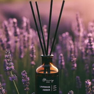 May include: A brown glass diffuser with black reeds, labelled "Lavender Amber." The diffuser is set on a wooden base, surrounded by purple lavender flowers. The background is blurred, creating a soft focus.