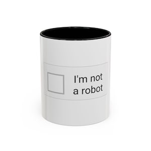 May include: A white ceramic mug with a black interior rim. The exterior features a rectangular graphic containing an empty square checkbox and the black text "I'm not a robot" printed in a sans-serif font on the side of the cylindrical vessel.
