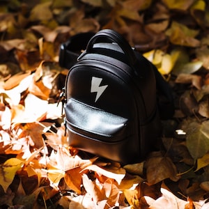 May include: Black leather backpack with a white lightning bolt design on the front. The small backpack has a top handle, a front pocket, and a zipper closure. The bag is sitting on a bed of fallen autumn leaves.