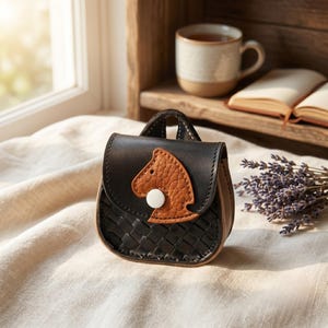 Op de afbeelding: Een kleine zwarte leren tas met een geweven textuur op de voorflap. De flap is voorzien van een lichtbruine, getextureerde applicatie in de vorm van een paard, vastgezet met een enkele witte ronde drukknoop. De tas heeft een handvat aan de bovenkant.
