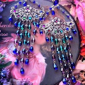 May include: A pair of silver-toned chandelier earrings featuring a top blue oval stone, a fan-shaped filigree centerpiece with small colorful crystals, and multiple dangling chains adorned with blue, purple, and teal teardrop-shaped beads.