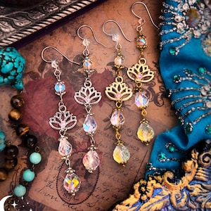 May include: Handmade Kerala Rose earrings with lotus flower and crystal drop details. The earrings are silver and gold-toned, with iridescent and clear crystal beads. The background includes a teal fabric, a candle, and other jewelry.