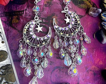 May include: A pair of silver-toned dangle earrings featuring a crescent moon frame with a small star charm. The moon is adorned with iridescent crystals and multiple hanging chains with faceted clear crystal beads in teardrop and round shapes.