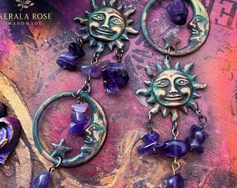 May include: A pair of dangle earrings featuring antique bronze-toned sun and crescent moon faces. Each earring incorporates raw, deep purple amethyst stone chips and small star charms, suspended from dark metal hooks with a weathered finish.