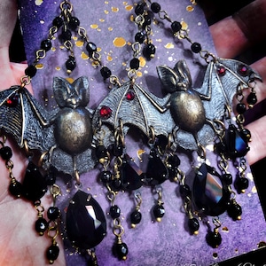 May include: A pair of dangle earrings featuring bronze-toned bat charms with small red crystal accents on the wings. The bats are suspended from bronze chains adorned with black faceted beads and large black teardrop-shaped crystal pendants.