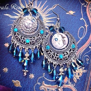 May include: A pair of silver-toned dangle earrings featuring a sun face on one and a crescent moon face on the other. The earrings have intricate filigree metalwork, small teal crystal accents, and multiple dangling dark blue teardrop beads.