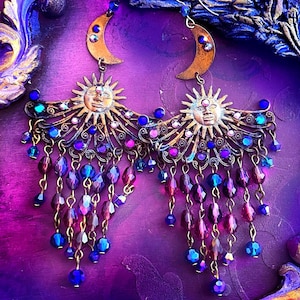 May include: A pair of dangle earrings featuring a brass-toned crescent moon and a sun face with radiating rays. The design is accented with small crystals and multiple hanging chains adorned with faceted purple and blue glass beads.