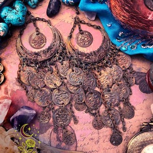 Può includere: Un paio di orecchini pendenti color bronzo caratterizzati da una montatura a mezzaluna con un ciondolo a moneta centrale e molteplici dischi metallici pendenti di forma circolare e a goccia. Gli orecchini hanno una finitura testurizzata.