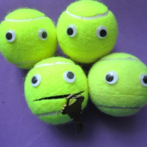 May include: Four bright neon yellow tennis balls arranged in a cluster. Each ball features two attached plastic googly eyes. One of the tennis balls has a horizontal slit cut into its surface, with a metallic key inserted into the opening.