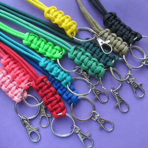 May include: A collection of colorful paracord keychains with silver key rings and clips. The keychains are made with different colors of paracord, including pink, red, blue, green, and yellow.