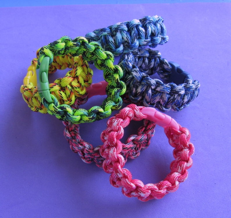 May include: Five colorful paracord bracelets with plastic clasps. The bracelets are in shades of blue, green, yellow, pink, and black.