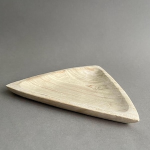 Può includere: Un vassoio triangolare in legno chiaro con un interno poco profondo e curvo. Il vassoio presenta un motivo a venature naturali del legno ed è posto su uno sfondo grigio neutro. La forma del vassoio è geometrica e moderna.