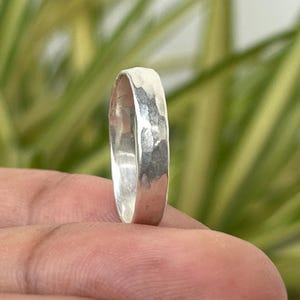 May include: A hammered silver ring with a textured surface. The ring is round and has a simple design. The ring is held up by a hand in front of a background of green and yellow leaves.