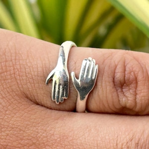 May include: A silver ring featuring two hands in an embracing design. One hand is open, and the other is reaching out to meet it. The ring is worn on a finger, with a blurred green and yellow background.