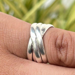 Könnte beinhalten: Ein silberner Ring mit einem geflochtenen Design. Der Ring besteht aus drei Bändern, die sich verschlingen und so einen strukturierten, auffälligen Look erzeugen. Der Ring wird an einem Finger gezeigt, mit einem verschwommenen grünen Hintergrund.