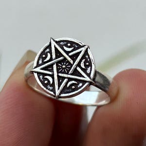 May include: A silver ring with a detailed pentagram design within a circular frame. The ring has a polished finish and is held up against a soft background. The pentagram is a five-pointed star, a symbol often associated with Wicca and Paganism.