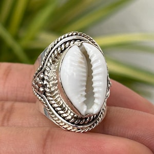 Puede incluir: Un anillo de plata con una concha de cauri blanca como pieza central. El anillo tiene una banda detallada y texturizada con un diseño trenzado alrededor de la concha. El anillo se muestra en una mano, con un fondo verde borroso.