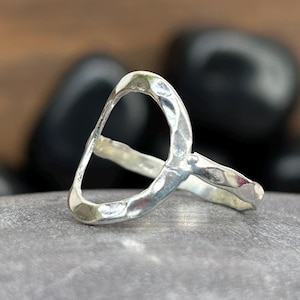 Op de afbeelding: Een gehamerde zilveren ring met een open, abstract ontwerp. De ring heeft een getextureerd oppervlak en een gladde band. Het wordt getoond op een grijze steen, met zwarte stenen en een houten achtergrond.