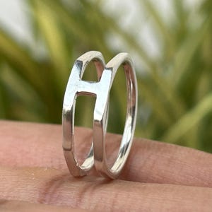 May include: A silver ring with the letter "A" as its main design element. The ring has a double band and the letter is open, giving it a modern and elegant appearance. The ring is displayed on a hand.