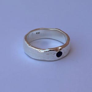 May include: A silver ring with a textured surface and a small black stone. The ring is a simple band design, with a slightly irregular shape. The ring is set against a light background.