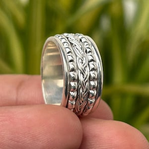 May include: A silver ring with a braided center design and beaded edges. The ring is held in a person's hand, with a blurred green background.
