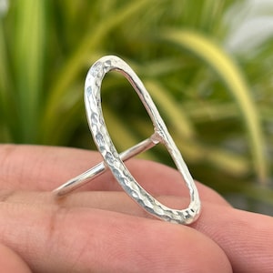 Pode incluir: Um anel de prata com um design oval alongado. O anel tem uma textura martelada e uma banda simples. O anel é mostrado em uma mão com um fundo verde desfocado.