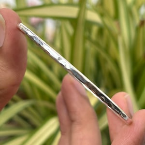 Könnte beinhalten: Ein gehämmertes Silberarmband. Das Armband hat eine strukturierte Oberfläche und ein schlichtes, minimalistisches Design. Das Silber reflektiert das Licht und erzeugt ein helles, glänzendes Aussehen. Das Armband wird in einer Hand vor einem Hintergrund aus grünem Laub gehalten.