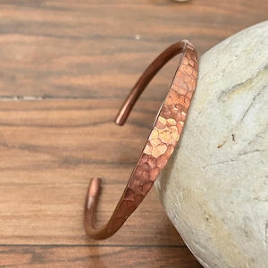 May include: A copper cuff bracelet with a hammered texture. The open-ended bracelet is a warm reddish-brown color and rests on a wooden surface next to a smooth, light-colored stone. The bracelet is approximately 2.5 inches in diameter.