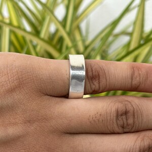 May include: A wide, polished silver ring. The ring is a simple band design, with a flat surface. The ring is worn on a finger, with a blurred background of green and yellow foliage.