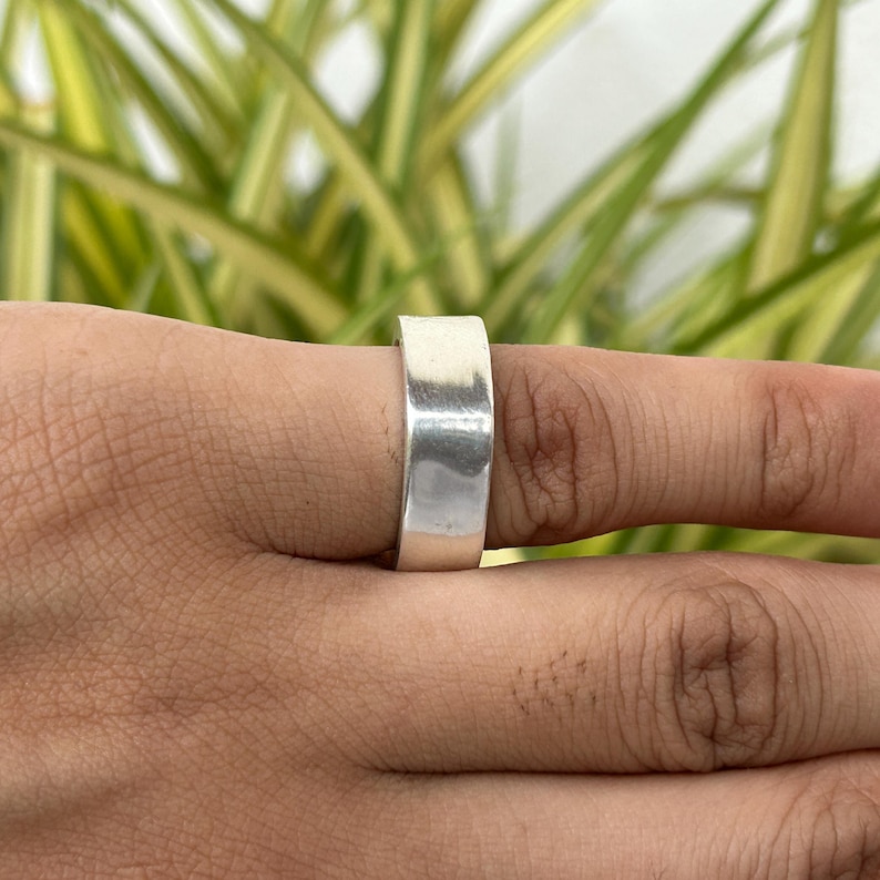 May include: A wide, polished silver ring. The ring is a simple band design, with a flat surface. The ring is worn on a finger, with a blurred background of green and yellow foliage.