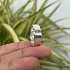 May include: A silver ring with a flat band. The ring is held between fingers against a background of green and yellow leaves. The ring has a polished, reflective surface.