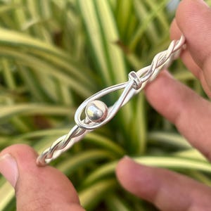May include: A silver-colored bangle bracelet with a twisted rope design. The bracelet features a small, round, silver bead held in place by a loop. The bracelet is held in front of a background of green and yellow leaves.