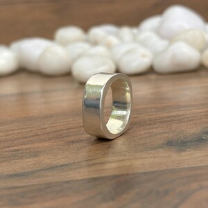 May include: A silver-colored ring with a square shape. The ring is made of a shiny metal and has a smooth surface. The ring is displayed on a wooden surface with white stones in the background.