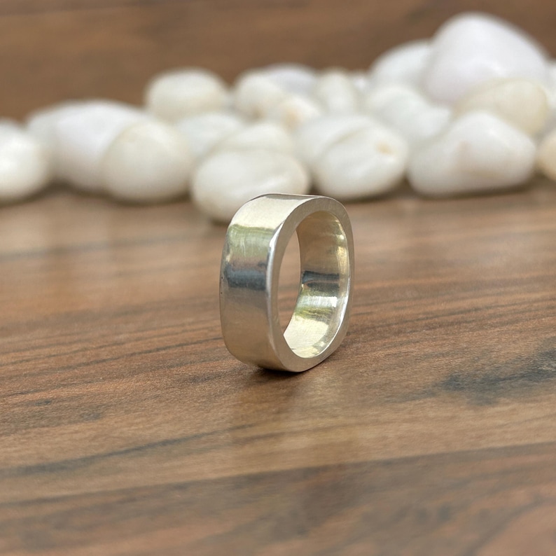 May include: A silver-colored ring with a square shape. The ring is made of a shiny metal and has a smooth surface. The ring is displayed on a wooden surface with white stones in the background.