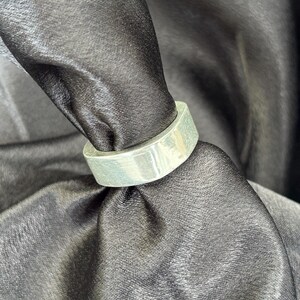 May include: A silver-colored ring with a flat, rectangular band. The ring is shown against a black, textured background, highlighting its metallic sheen. The ring's design is simple and modern, suitable for everyday wear.