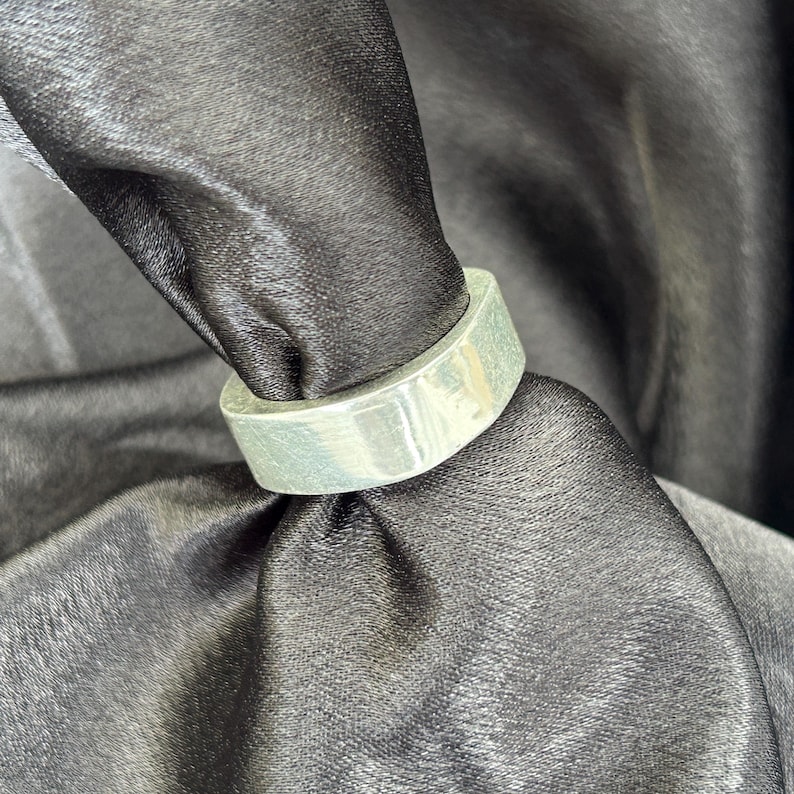 May include: A silver-colored ring with a flat, rectangular band. The ring is shown against a black, textured background, highlighting its metallic sheen. The ring's design is simple and modern, suitable for everyday wear.