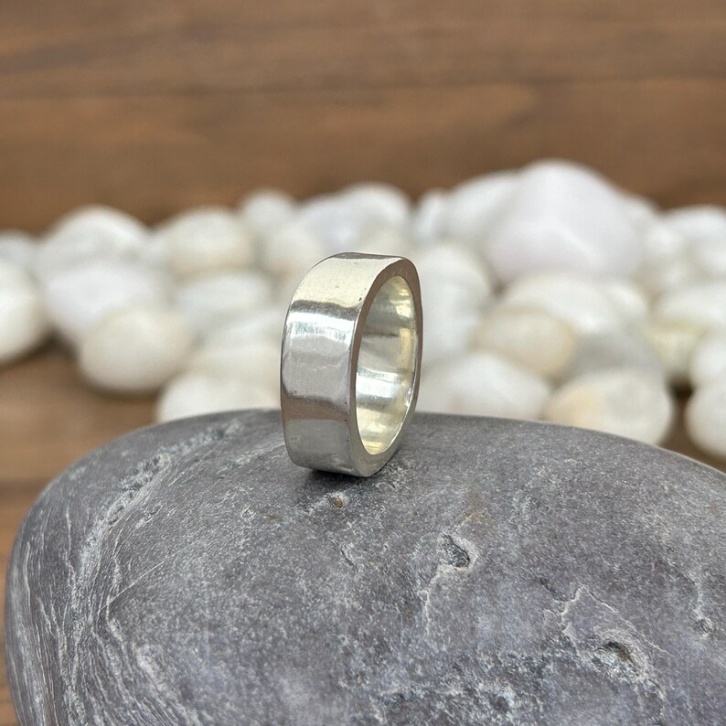 May include: A silver ring with a flat, rectangular design. The ring is smooth and reflective, with a polished finish. The ring is set against a background of white stones and a gray rock.