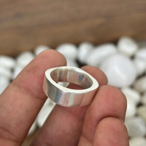 May include: A silver ring with a flat, rectangular band and rounded corners. The ring has a brushed metal finish and is held in a hand. The ring is a simple, modern design.