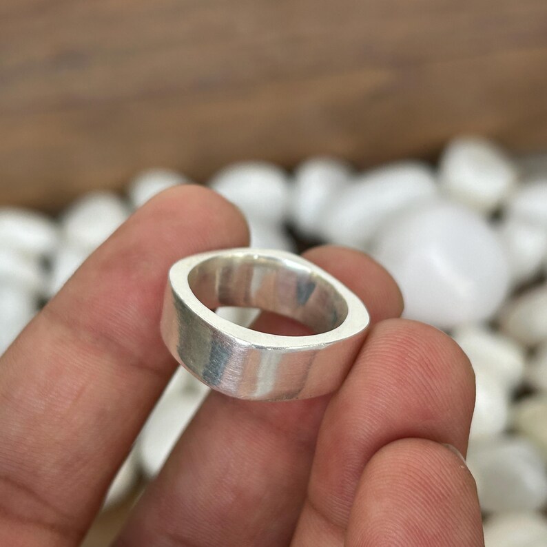 May include: A silver ring with a flat, rectangular band and rounded corners. The ring has a brushed metal finish and is held in a hand. The ring is a simple, modern design.