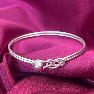 May include: A silver bangle bracelet featuring a knot design and a small ball clasp. The bracelet is made of two parallel bands and rests on a deep pink satin fabric. The jewellery is simple and elegant.