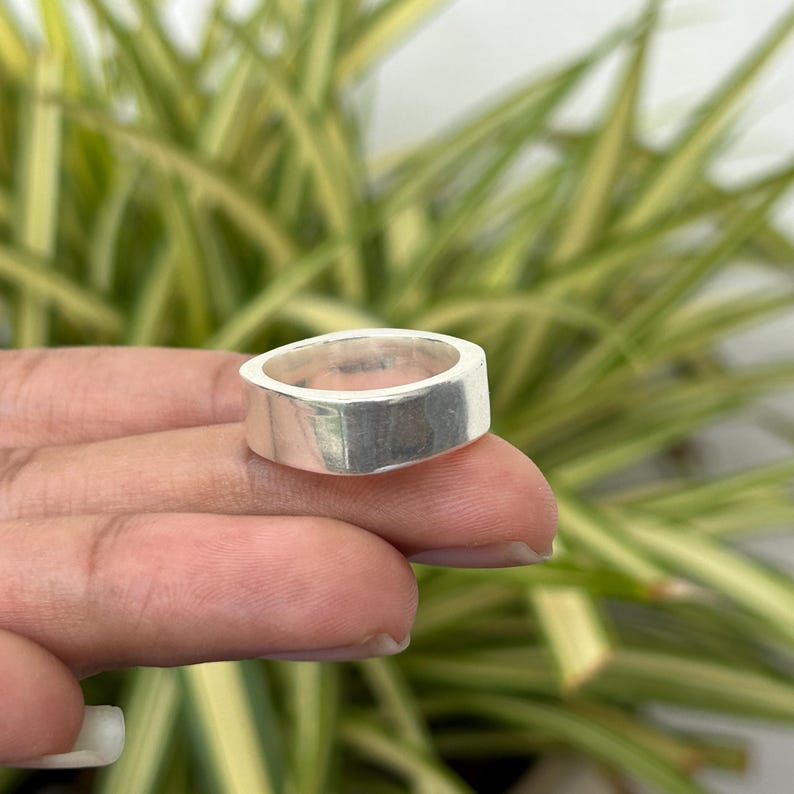 May include: A silver ring with a rectangular band. The ring is held up by a person's fingers. The background is a blurred view of green and yellow leaves. The ring is a simple, modern design.